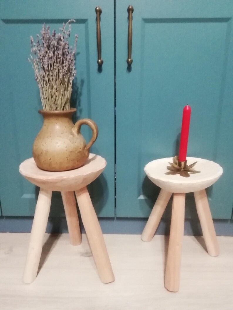 May include: Two wooden stools with three legs each. The stool on the left has a light brown top and a brown ceramic vase with dried lavender on top. The stool on the right has a white top and a red candle on top.