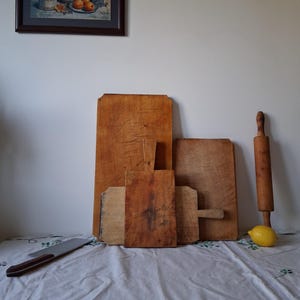Op de afbeelding: Een verzameling van vier rechthoekige houten snijplanken van verschillende groottes met zichtbare slijtage, een houten deegroller, een metalen mes met een bruin handvat en een gele citroen op een witte stoffen ondergrond.