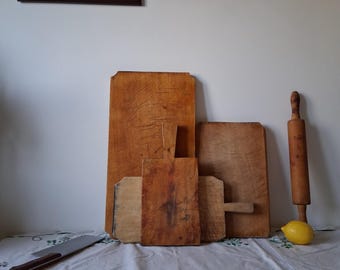 May include: A collection of four rectangular wooden cutting boards of varying sizes with visible surface wear, a wooden rolling pin, a metal knife with a brown handle, and a single yellow lemon arranged together on a white fabric surface.