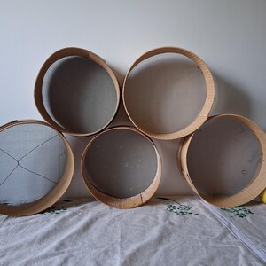 May include: Five round wooden sifters with mesh bottoms, arranged in a semi-circle on a white fabric surface. A yellow lemon sits on the fabric to the right of the sifters.