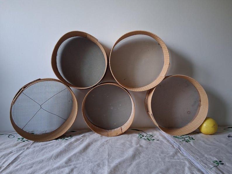 May include: Five round wooden sifters with mesh bottoms, arranged in a semi-circle on a white fabric surface. A yellow lemon sits on the fabric to the right of the sifters.