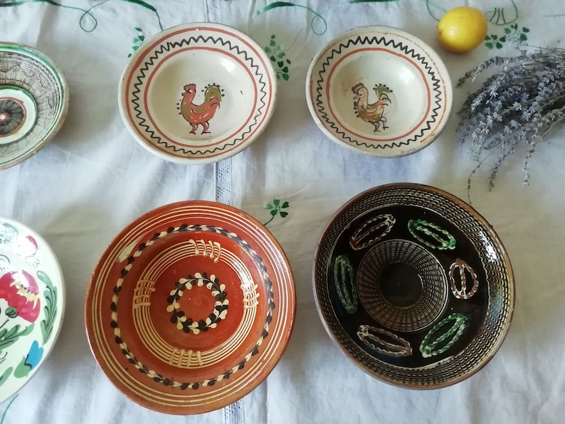 May include: Five ceramic bowls with different designs. The bowls are white, brown, and black with painted details. The designs include a rooster, a wreath, and geometric patterns.