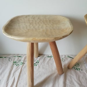 May include: A wooden stool with a wide, carved seat and three sturdy legs. The stool is made of light-colored wood and has a rustic, handmade appearance.