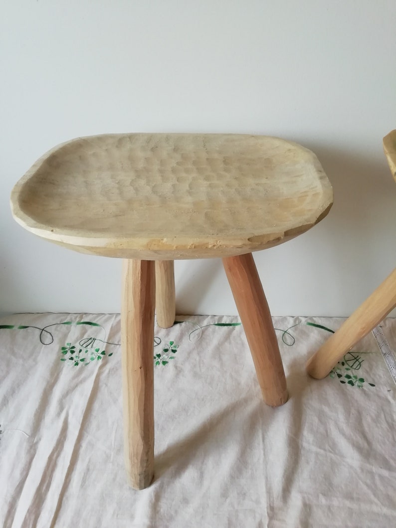 May include: A wooden stool with a wide, carved seat and three sturdy legs. The stool is made of light-colored wood and has a rustic, handmade appearance.