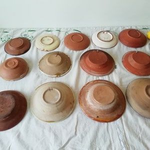 May include: A collection of 12 terracotta and glazed ceramic plates with various designs. The plates are arranged in two rows on a white tablecloth. Some of the plates have a brown glaze, while others have a white glaze with floral designs.