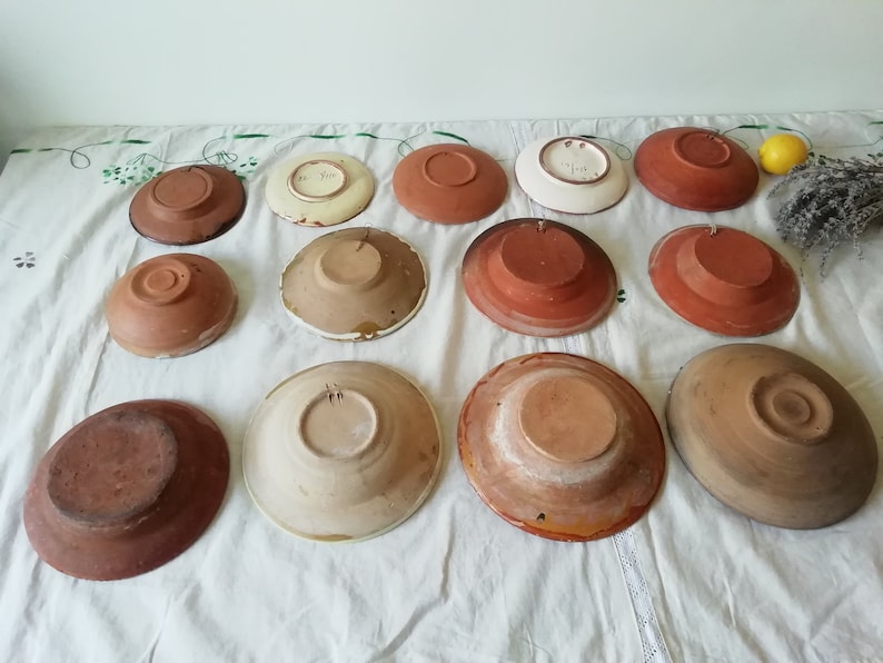 May include: A collection of 12 terracotta and glazed ceramic plates with various designs. The plates are arranged in two rows on a white tablecloth. Some of the plates have a brown glaze, while others have a white glaze with floral designs.
