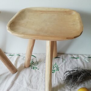 May include: A wooden stool with a wide, flat top and three legs. The stool is made of natural wood and has a rustic, handmade look.