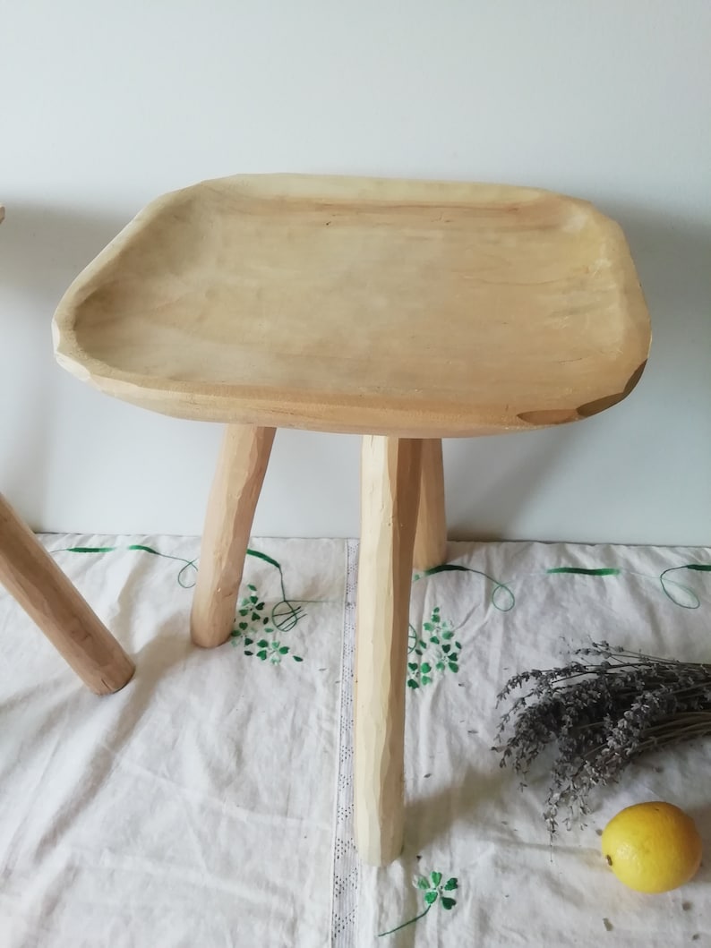 May include: A wooden stool with a wide, flat top and three legs. The stool is made of natural wood and has a rustic, handmade look.