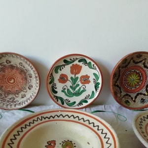 May include: Five ceramic plates with hand-painted designs. The plates are in various sizes and feature different patterns, including floral motifs, geometric shapes, and swirls. The plates are all in shades of brown, orange, green, and white.