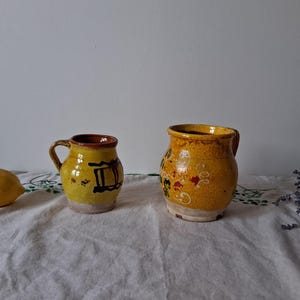 Peut inclure: Deux pichets en céramique jaune avec des motifs peints en brun et blanc. Le plus petit pichet a une anse et le plus grand pichet a une bouche plus large.