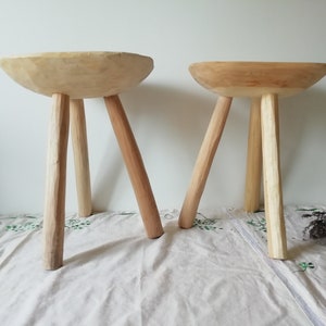 May include: Two wooden stools with a bowl-shaped seat and three legs. The stools are made of natural wood and have a rustic, handmade look.