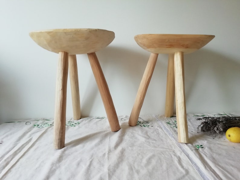 May include: Two wooden stools with a bowl-shaped seat and three legs. The stools are made of natural wood and have a rustic, handmade look.