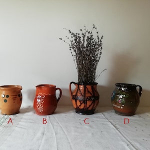 以下が含まれることがあります： 異なるペイントデザインが施された、茶色の陶器のピッチャーが4つあります。中央のピッチャーは、乾燥したラベンダーで満たされており、黒い釉薬がかけられています。