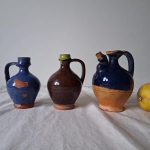 Può includere: Tre piccole caraffe in ceramica con smalto blu, marrone e arancio. Le caraffe hanno un manico e un beccuccio. Le caraffe sono appoggiate su una tovaglia bianca.