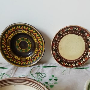 May include: Four ceramic plates with hand-painted designs. The plates have brown, green, yellow, and orange accents. The plates are on a white tablecloth with green embroidery.
