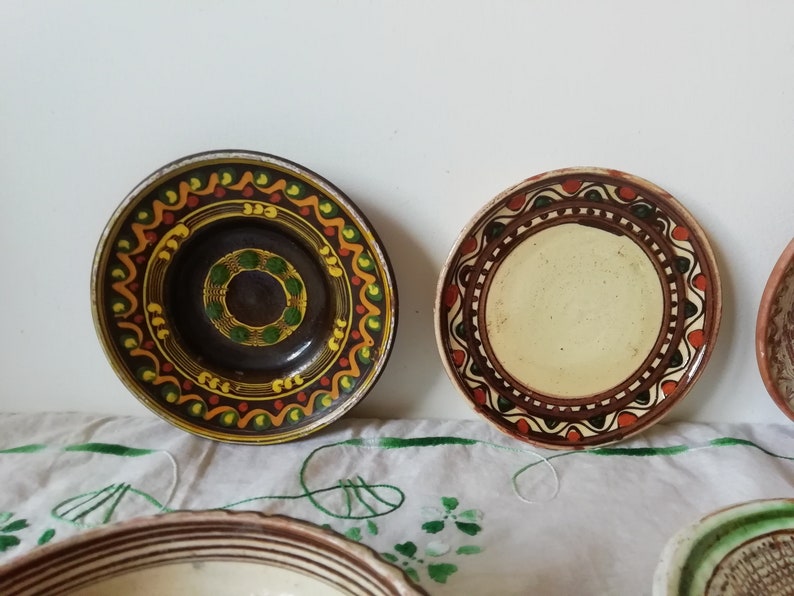 May include: Four ceramic plates with hand-painted designs. The plates have brown, green, yellow, and orange accents. The plates are on a white tablecloth with green embroidery.