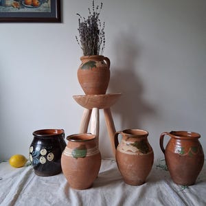 Könnte beinhalten: Vier Terrakotta-Krüge mit bemalten Designs. Die Krüge haben ein rustikales, handgemachtes Aussehen und werden auf einem Holzständer mit getrockneter Lavendel in einem ähnlichen Krug ausgestellt.