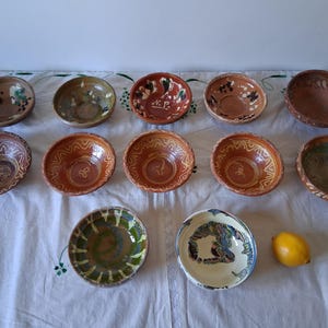Peut inclure: Un ensemble de douze bols en céramique disposés sur trois rangées, présentant des glaçures aux tons terreux, des motifs géométriques peints à la main et des motifs floraux. Un bol porte les lettres N.P. Un citron jaune est posé à côté.