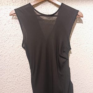 May include: A sleeveless black top featuring a deep V-neckline and a sheer, lightweight fabric construction. The garment is displayed on a wooden hanger, showcasing a simple, solid-coloured design with a slightly gathered side detail.