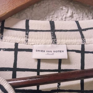 May include: Close-up of a cream-colored shirt with a black grid pattern. The label reads "DRIES VAN NOTEN Small". The shirt is hanging on a wooden hanger.