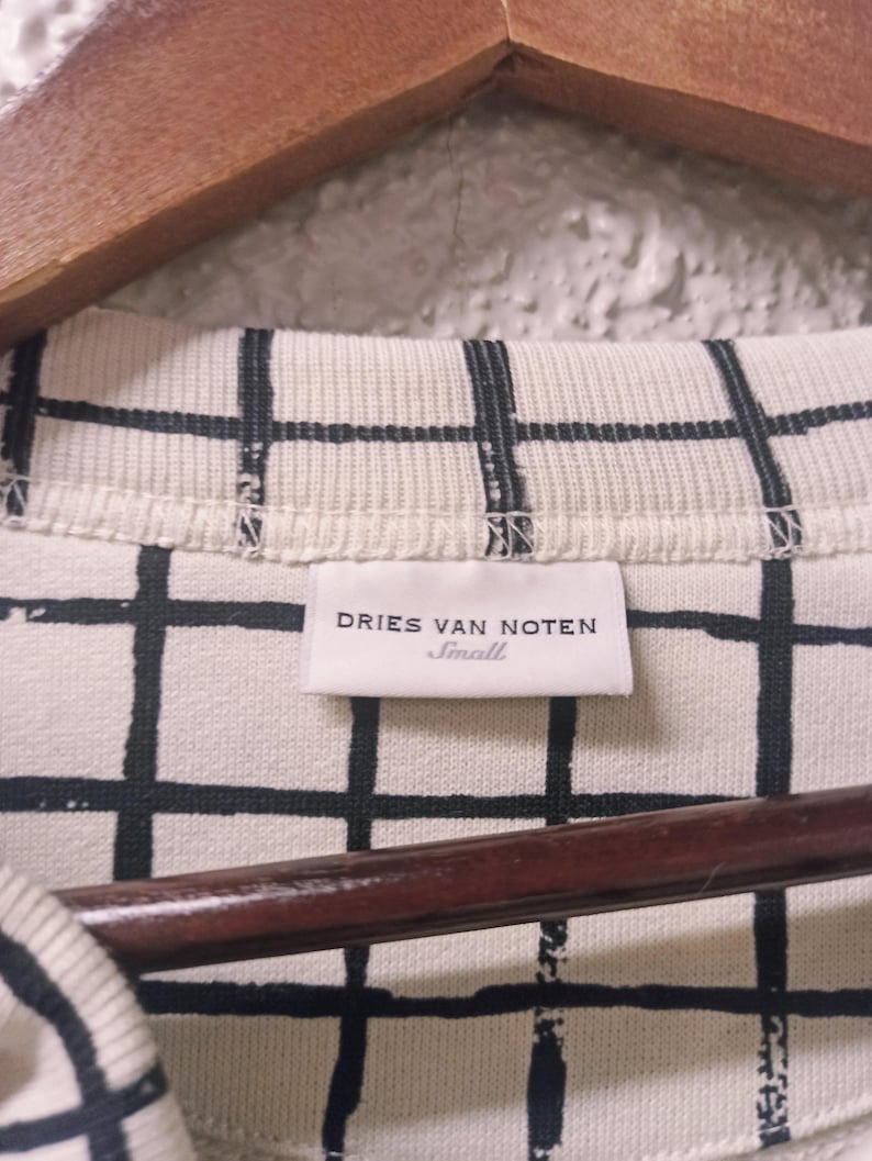 May include: Close-up of a cream-colored shirt with a black grid pattern. The label reads "DRIES VAN NOTEN Small". The shirt is hanging on a wooden hanger.