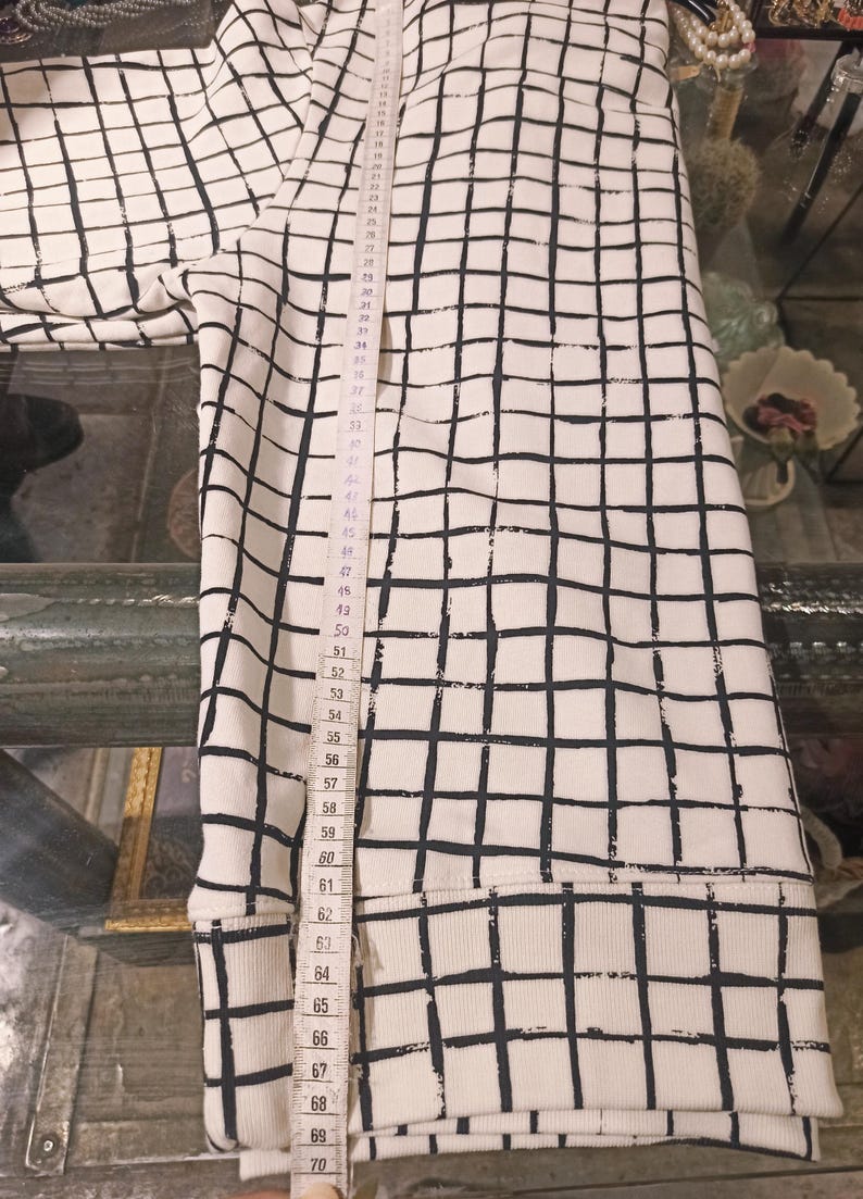 May include: White sweatshirt with a black grid pattern. The sweatshirt is measured with a tape measure, showing a length of approximately 70 inches. The garment has a ribbed hem and cuffs.