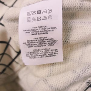May include: Close-up of a white clothing tag with care instructions and the text "100% COTTON". The tag also includes washing symbols and the phrase "MADE IN TURKEY". The background is a white and black patterned fabric.