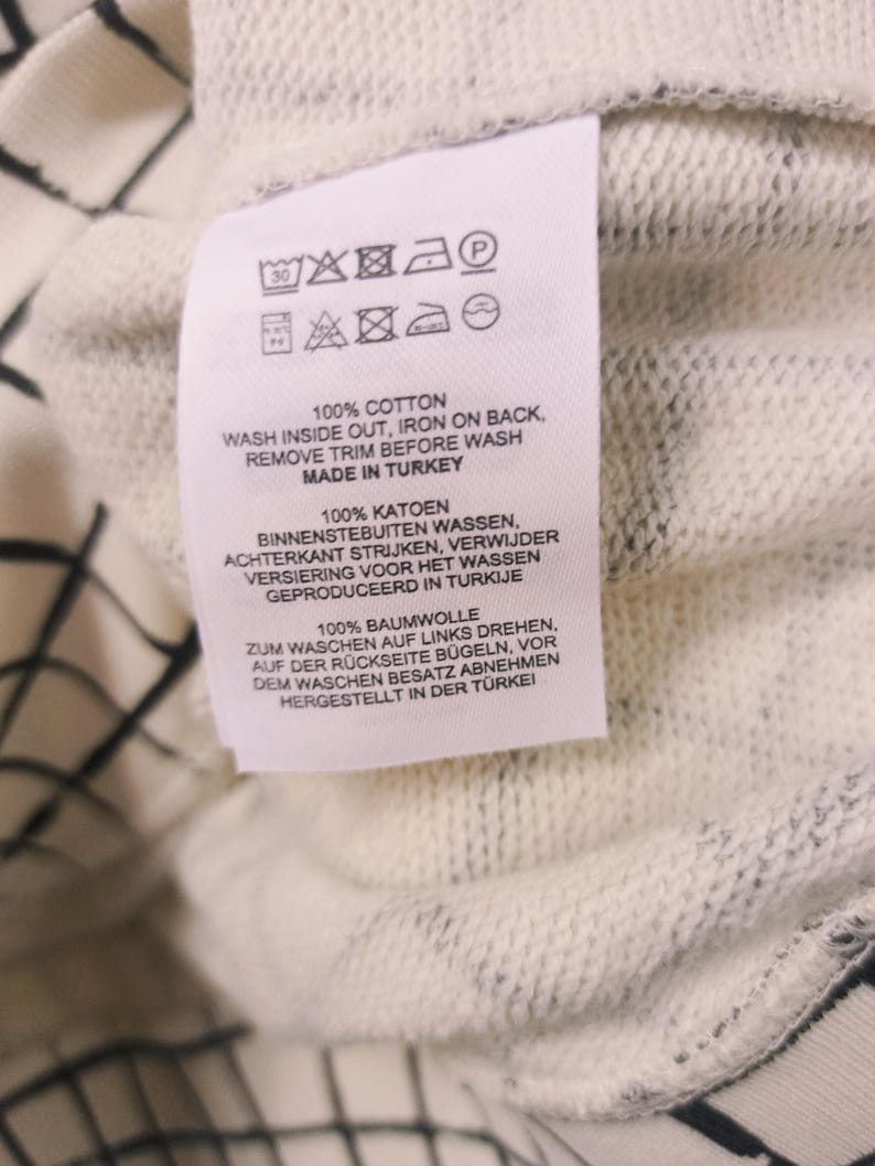 May include: Close-up of a white clothing tag with care instructions and the text "100% COTTON". The tag also includes washing symbols and the phrase "MADE IN TURKEY". The background is a white and black patterned fabric.