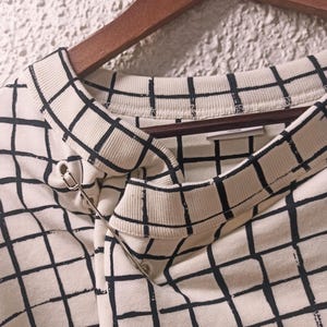 May include: Cream-colored sweatshirt with a black grid pattern, hanging on a wooden hanger. A silver safety pin secures the fabric at the neckline. The design is a modern take on a classic pattern.