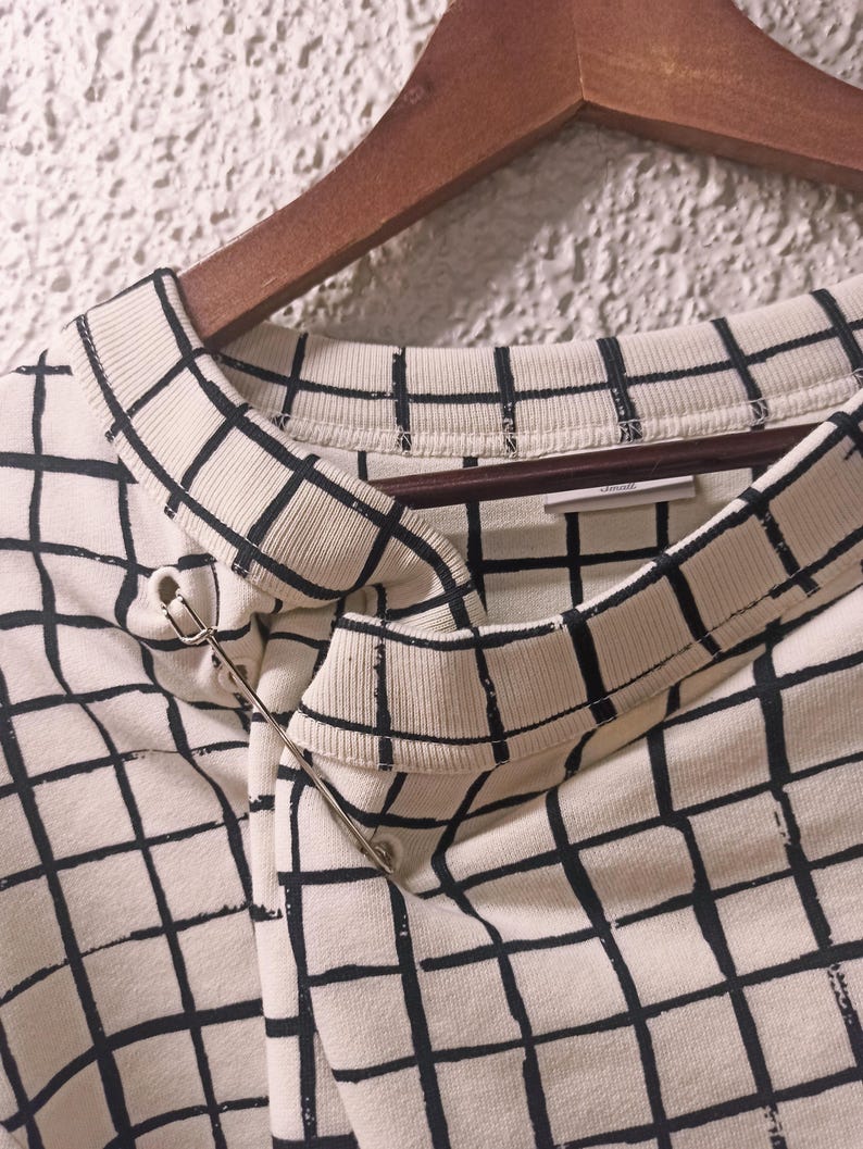 May include: Cream-colored sweatshirt with a black grid pattern, hanging on a wooden hanger. A silver safety pin secures the fabric at the neckline. The design is a modern take on a classic pattern.