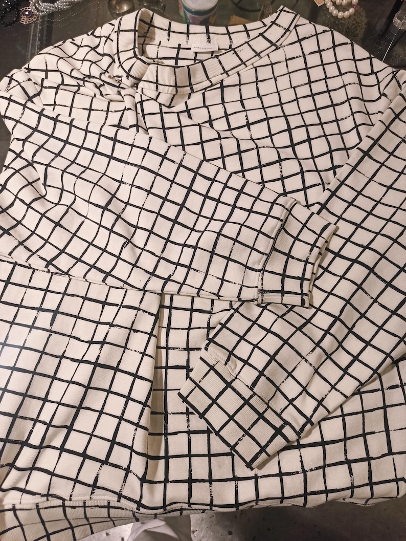 May include: Cream-colored sweatshirt with a black grid pattern. The long-sleeved top features a crew neck and ribbed cuffs. The grid design covers the entire garment, creating a modern, geometric look.