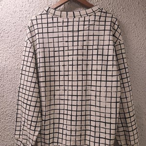 May include: Cream-colored sweatshirt with a black grid pattern. The long-sleeved top is hanging on a wooden hanger against a textured wall. The grid pattern covers the entire sweatshirt, including the sleeves and the body.