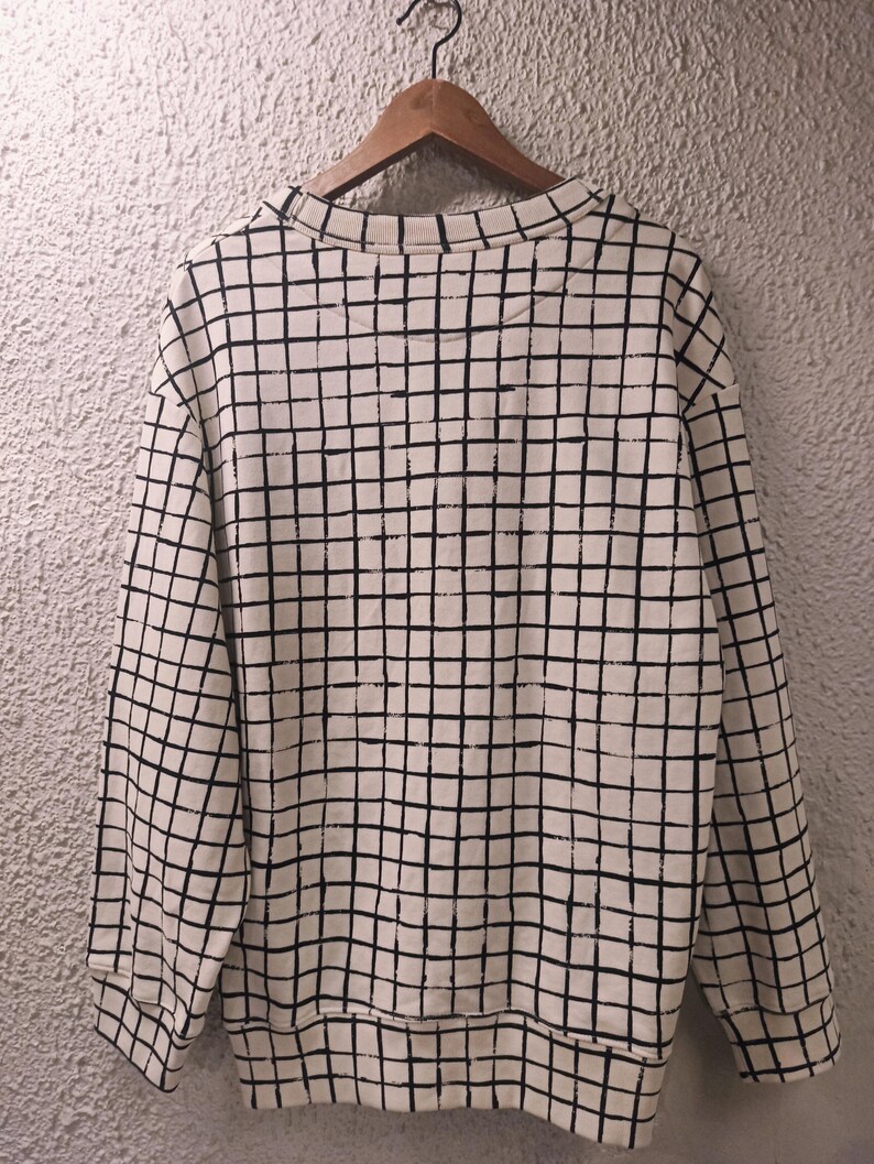 May include: Cream-colored sweatshirt with a black grid pattern. The long-sleeved top is hanging on a wooden hanger against a textured wall. The grid pattern covers the entire sweatshirt, including the sleeves and the body.
