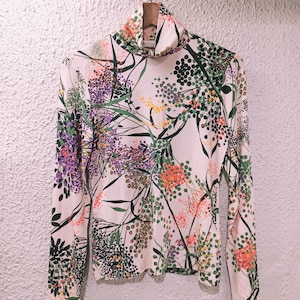 May include: A long-sleeved turtleneck shirt with a floral pattern. The shirt features a cream-colored base with a colorful botanical design in shades of purple, green, orange, and yellow. The shirt is hanging on a wooden hanger.