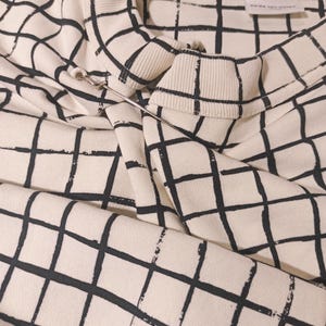May include: Cream-colored sweatshirt with a black grid pattern. The sweatshirt has a crew neck and long sleeves. A silver safety pin is attached to the collar. The tag reads "DRIES VAN NOTEN".