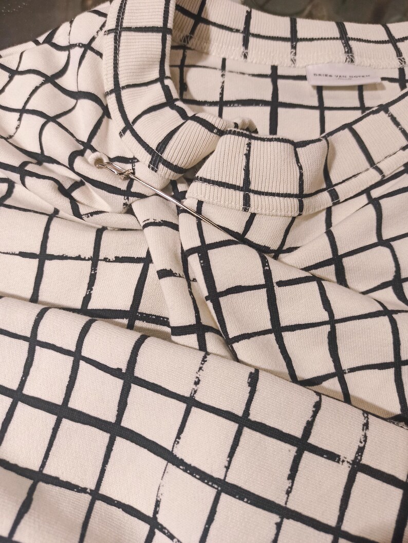 May include: Cream-colored sweatshirt with a black grid pattern. The sweatshirt has a crew neck and long sleeves. A silver safety pin is attached to the collar. The tag reads "DRIES VAN NOTEN".
