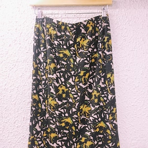May include: A knee-length skirt with an elasticated waistband, displaying a floral pattern in yellow, green, and black on a cream backdrop. The skirt is suspended on a wooden hanger against a white wall.