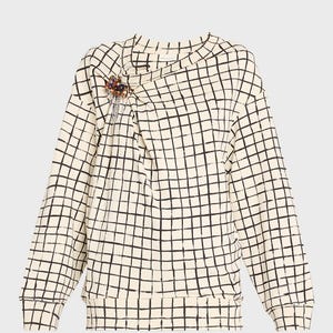 May include: Cream-colored long-sleeved sweatshirt with a black grid pattern. The neckline has a draped design, and a decorative brooch with colorful beads is pinned on the left side. The sweatshirt has a ribbed hem and cuffs.