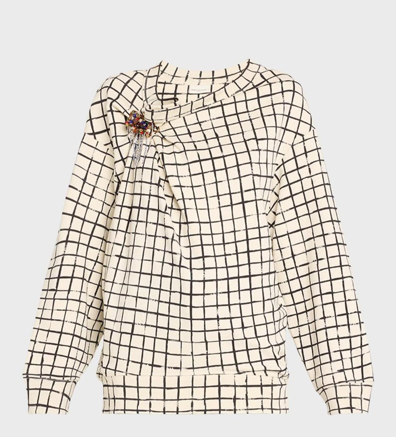 May include: Cream-colored long-sleeved sweatshirt with a black grid pattern. The neckline has a draped design, and a decorative brooch with colorful beads is pinned on the left side. The sweatshirt has a ribbed hem and cuffs.