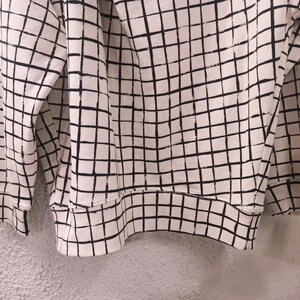 May include: Cream-colored sweatshirt with a black grid pattern. The long-sleeved top features a ribbed hem and cuffs. The grid pattern covers the entire garment, creating a geometric design.