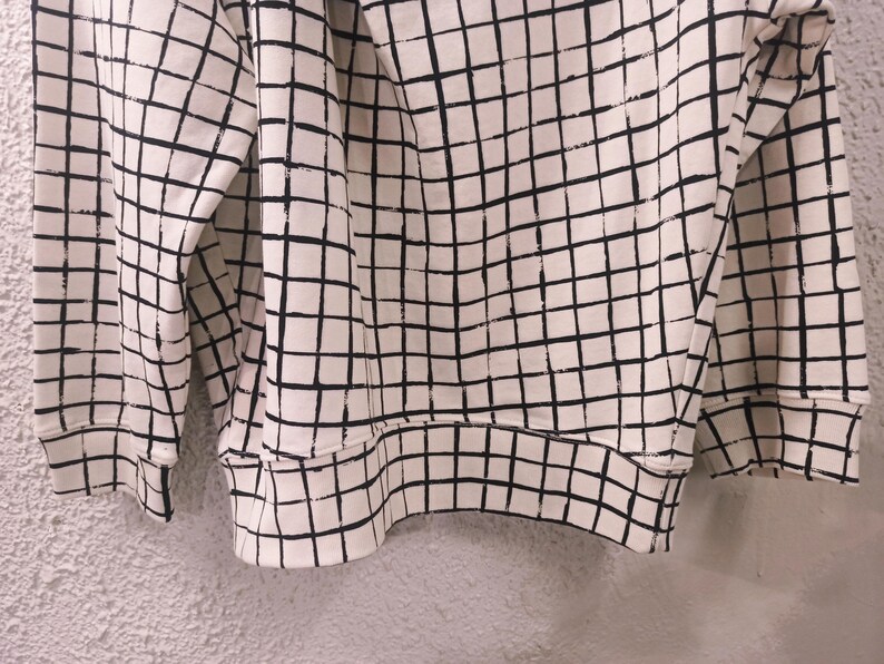 May include: Cream-colored sweatshirt with a black grid pattern. The long-sleeved top features a ribbed hem and cuffs. The grid pattern covers the entire garment, creating a geometric design.