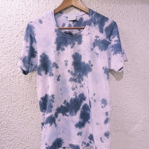 Peut inclure: Un t-shirt à manches courtes et col rond, orné d'un motif tie-dye bleu et blanc. Le t-shirt est suspendu à un cintre en bois. Le motif tie-dye présente des formes irrégulières en forme de nuages sur un mur neutre.