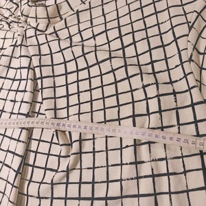 May include: A cream-colored long-sleeved shirt with a black grid pattern. A measuring tape is laid across the shirt, showing measurements in inches. The shirt's design is a repeating pattern of black lines on a cream background.