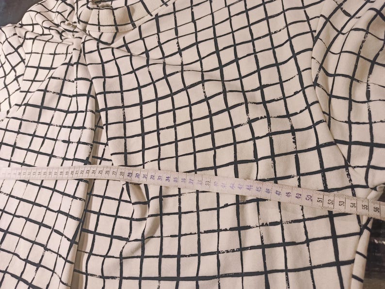 May include: A cream-colored long-sleeved shirt with a black grid pattern. A measuring tape is laid across the shirt, showing measurements in inches. The shirt's design is a repeating pattern of black lines on a cream background.