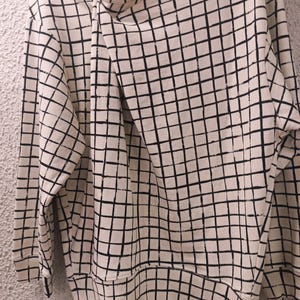 May include: Cream-colored sweatshirt with a black grid pattern. The long-sleeved top features a unique asymmetrical neckline with a button closure. The grid pattern covers the entire garment, including the sleeves and body.