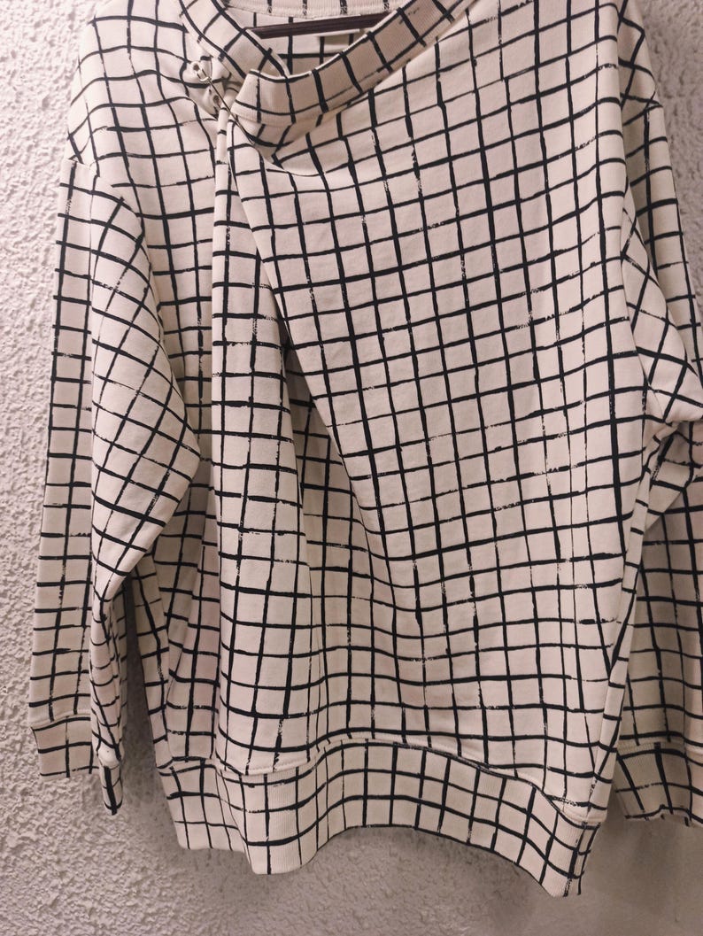 May include: Cream-colored sweatshirt with a black grid pattern. The long-sleeved top features a unique asymmetrical neckline with a button closure. The grid pattern covers the entire garment, including the sleeves and body.