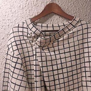 May include: Cream-colored long-sleeved shirt with a black grid pattern. The shirt is hanging on a wooden hanger. A silver pin secures the neckline. The fabric appears to be a soft, comfortable material.