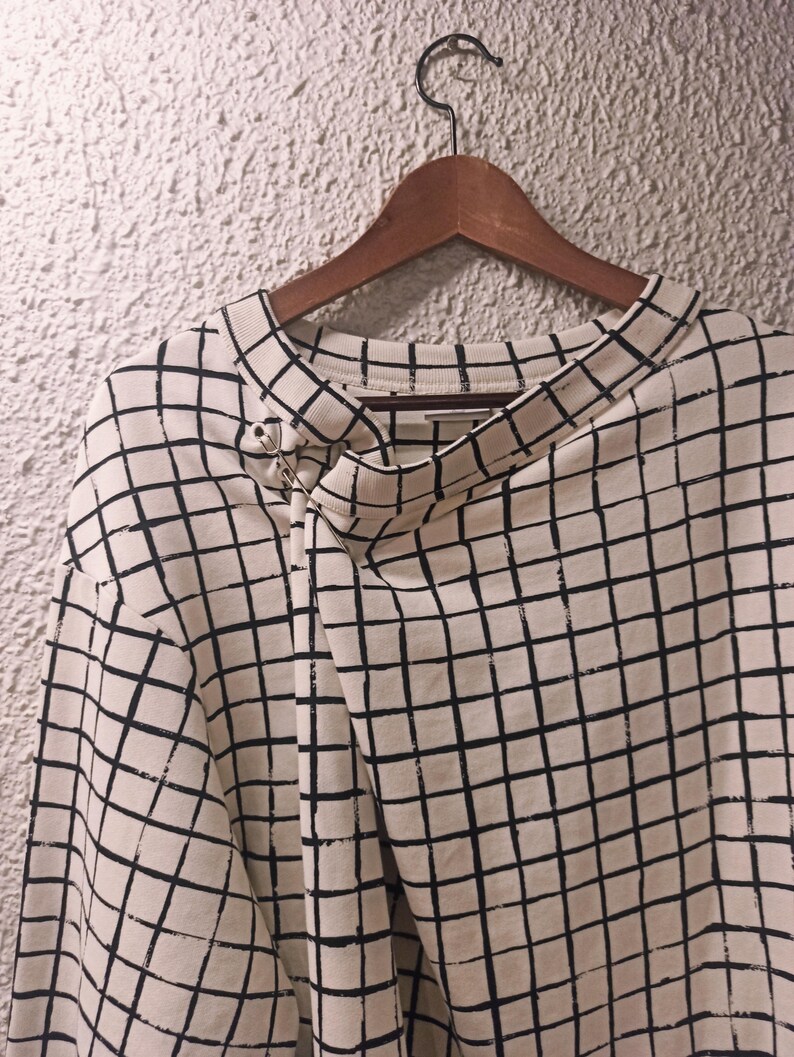 May include: Cream-colored long-sleeved shirt with a black grid pattern. The shirt is hanging on a wooden hanger. A silver pin secures the neckline. The fabric appears to be a soft, comfortable material.