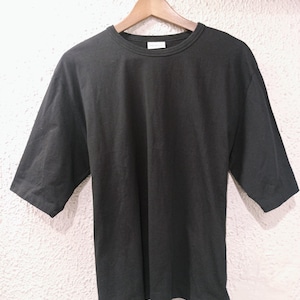 May include: A black, short-sleeved t-shirt hangs from a wooden hanger. The shirt has a crew neck and a relaxed fit. The sleeves extend to the elbows. The background is a textured, off-white wall.