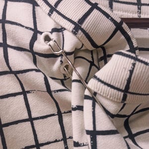 May include: Close-up of a cream-colored sweater with a black grid pattern. A silver safety pin is attached to the collar. The sweater is hanging on a wooden hanger.